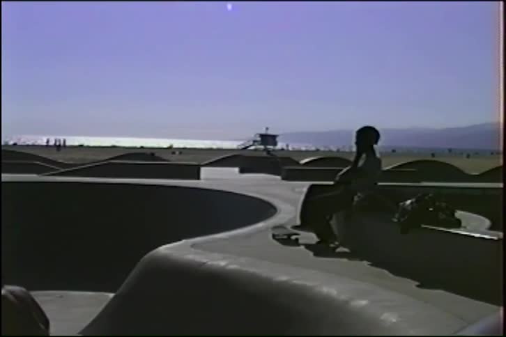 Skaters are silhouetted against the bright sun as they ride and relax at the Venice Beach skatepark in this vintage footage