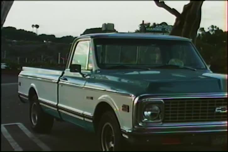 A classic two-tone teal and white Chevrolet C10 pickup truck parked in a lot in the 1980s