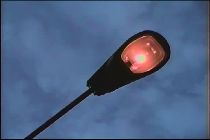 A street lamp flickers on, its reddish glow slowly illuminating against a dark and moody evening sky in vintage footage