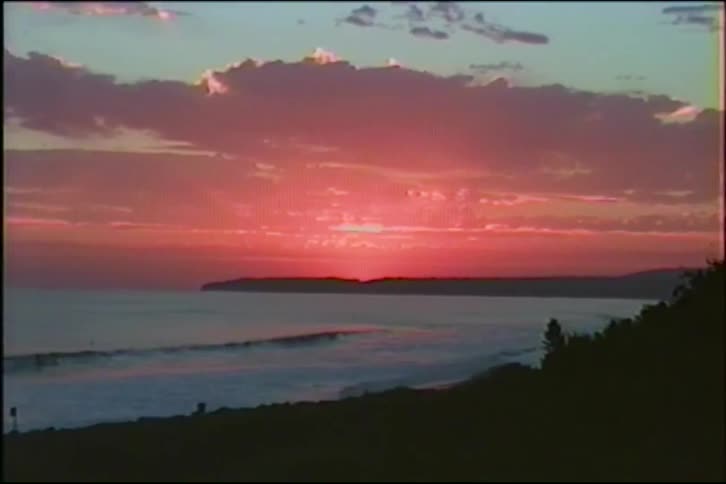 A vibrant pink and red sun sets behind the silhouette of distant hills over the ocean as waves gently roll to shore
