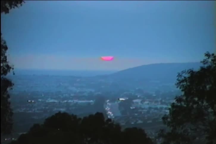Vintage VHS footage shows a surreal, vibrant pink sun setting behind mountains over a hazy city at dusk