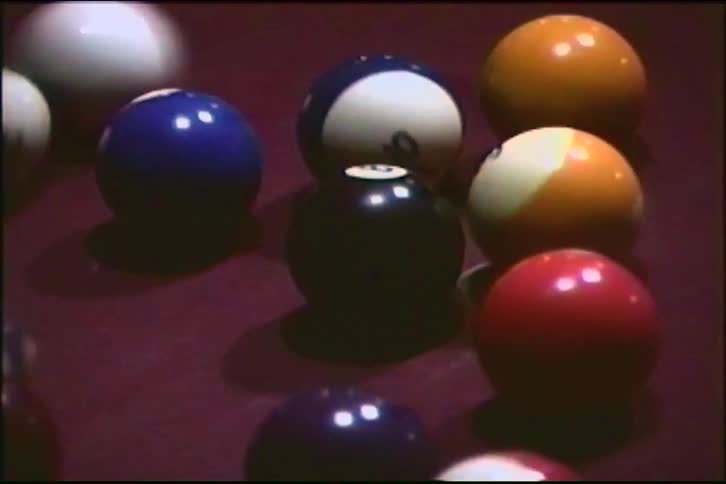 A close-up of a cue ball breaking a rack of colorful pool balls on a purple felt table in vintage-style footage