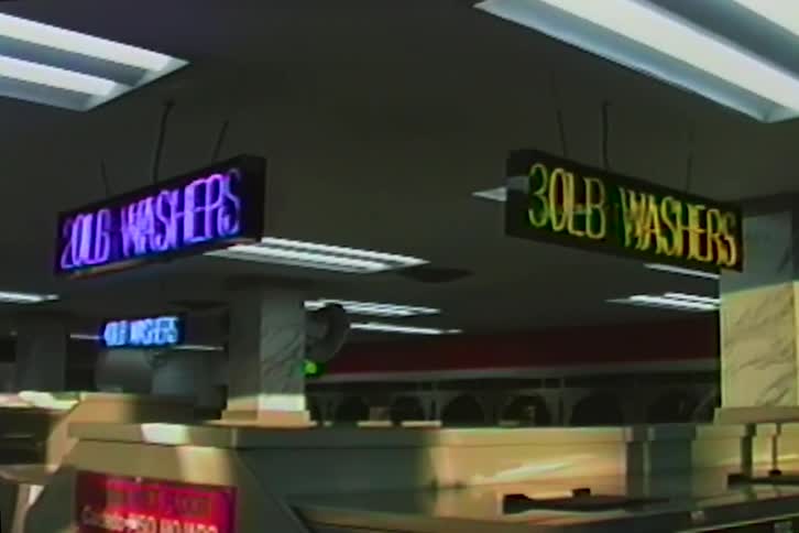 Colorful vintage light-up signs hang above rows of machines in a classic laundromat
