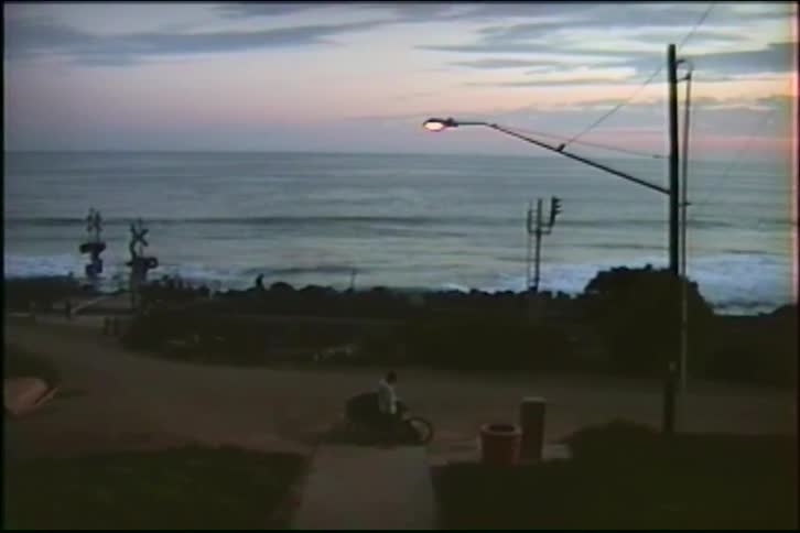 A lone cyclist watches the ocean waves from a coastal path next to a railroad crossing at dusk