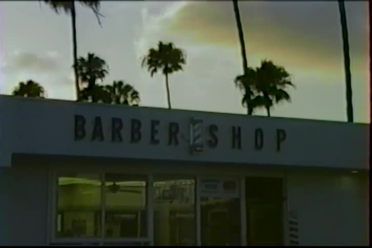 A vintage-style shot shows the exterior of a barbershop building with palm trees silhouetted against the sunset sky