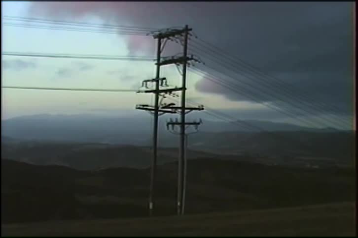 A slow zoom on two large power poles in a hilly rural landscape under a dark, moody sky at dusk