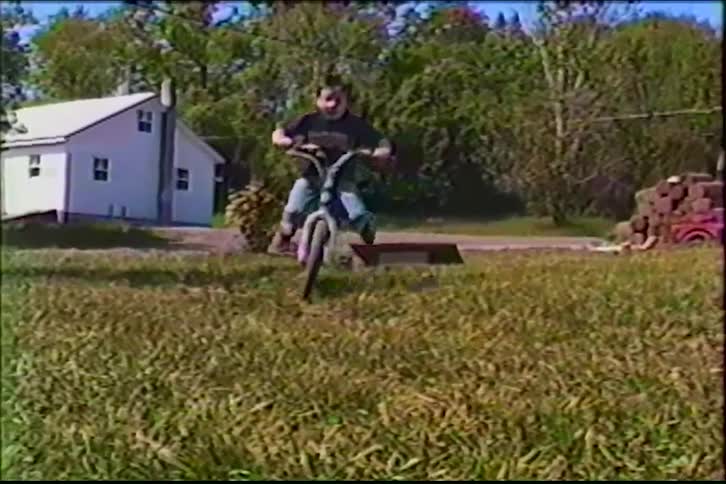 Vintage home video footage shows a young boy jumping his bicycle off a small ramp in a grassy backyard