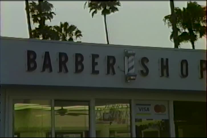 A slow pan reveals the vintage sign of a local barbershop with a spinning barber pole, set against a backdrop of palm trees at dusk