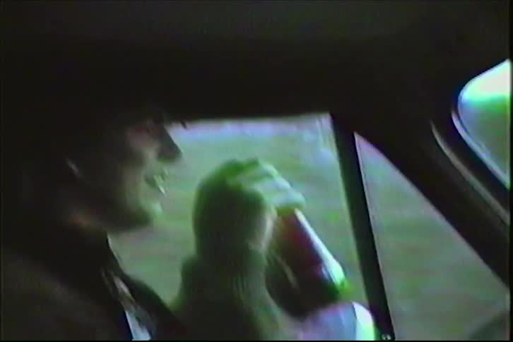 Grainy vintage footage captures a young man drinking a soda from a can while driving a car