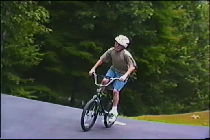 A young boy wearing a helmet energetically rides his BMX bike down the driveway in this vintage home video