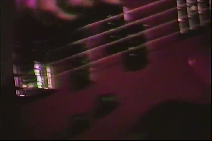 Vintage VHS footage zooms in on a rock musician's hands as he plays a red electric bass guitar on a dark stage