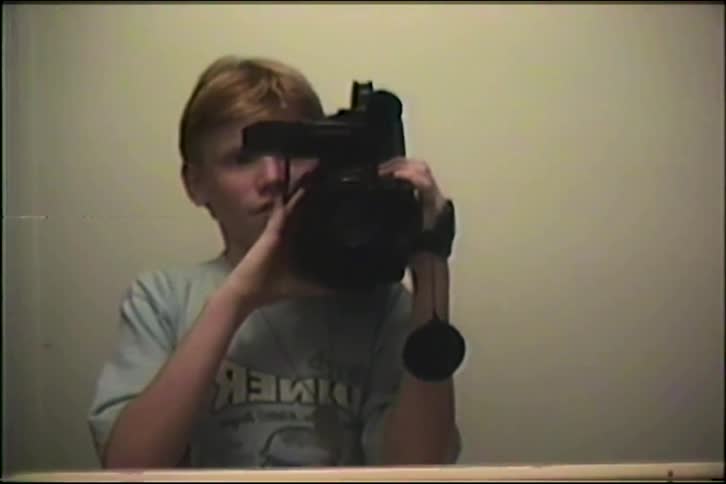 A teenage boy films his reflection in a mirror with a vintage camcorder, adjusting the camera as the lights slowly dim