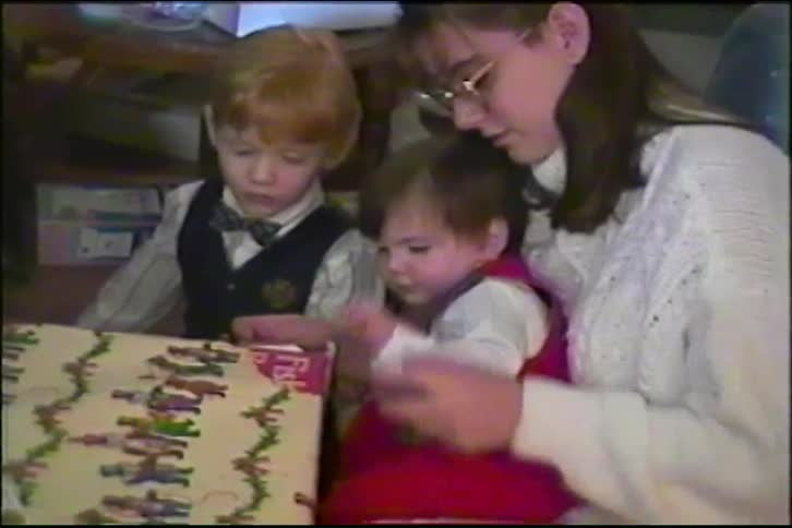 Vintage VHS footage shows a mother helping her young son and toddler daughter unwrap a Christmas gift