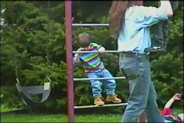A father with long hair lifts his daughter onto the monkey bars while his sons play nearby in a nostalgic home video from the 1990s