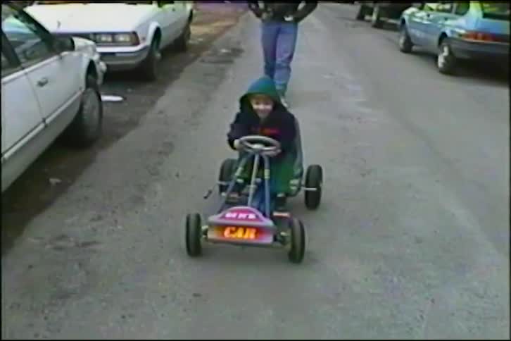 A father pushes his happy young son in a pedal go-kart down a residential street in this heartwarming vintage home video