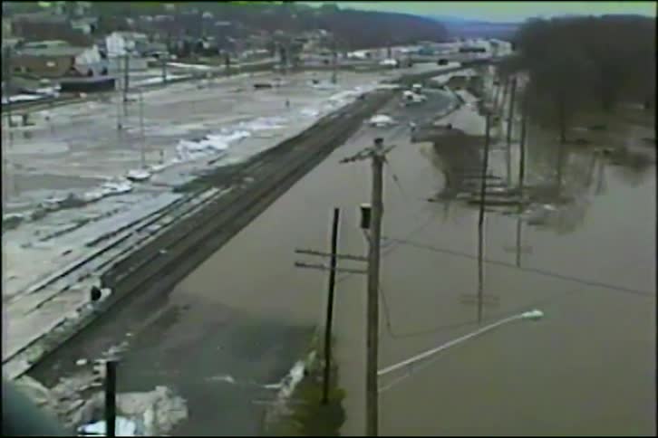 A wide archival shot captures severe flooding that has submerged railroad tracks, roads, and utility poles in a town
