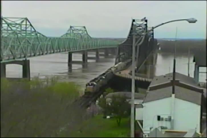 A vintage VHS shot captures a long freight train crossing a railroad bridge over a wide river on an overcast day