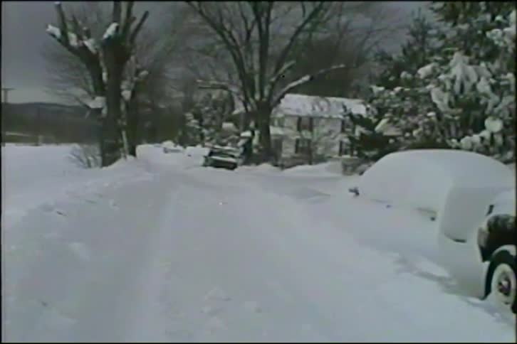 A vintage shot shows cars buried in deep snow on a residential street as a freight train passes by in the background