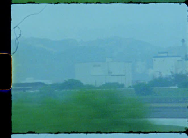 Grainy 8mm style footage shows a fast-moving, blurry view of a rural and industrial landscape with hazy mountains in the background