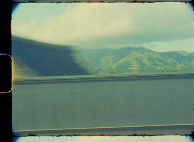 A dreamy, vintage 8mm film view of the Japanese countryside passing by a train window, showcasing lush green fields and distant mountains