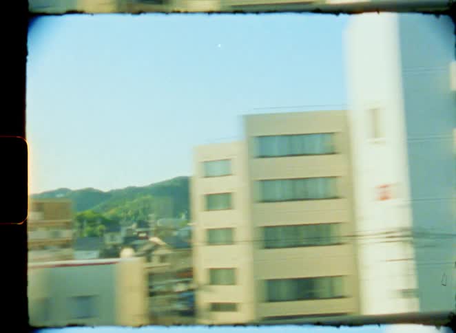 Vintage 8mm film captures a nostalgic view of a Japanese city with green hills in the background from a moving train window