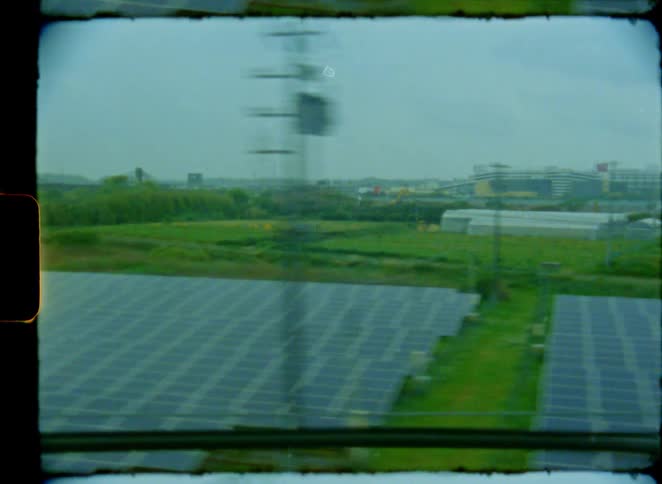 A vintage 8mm film captures the view from a moving train window, passing by industrial buildings, green fields, and a solar panel farm on an overcast day
