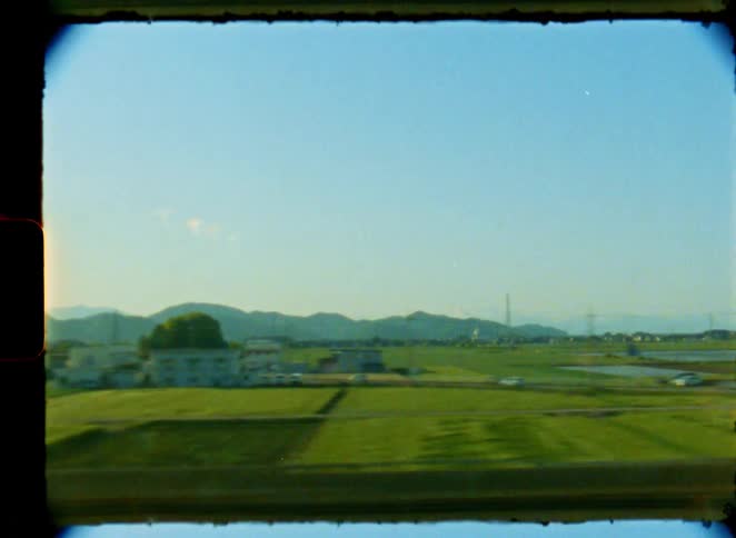 A scenic view of the Japanese countryside with rice paddies and mountains passing by from a train window, captured on vintage 8mm film