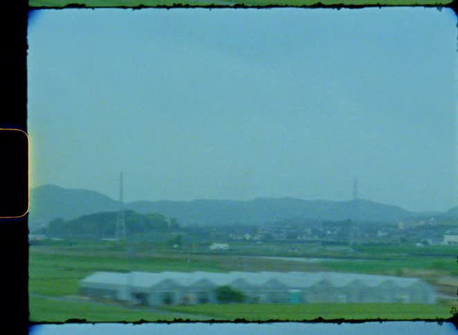 A vintage 8mm film captures the view of a rural landscape with green fields and distant mountains from a moving train window