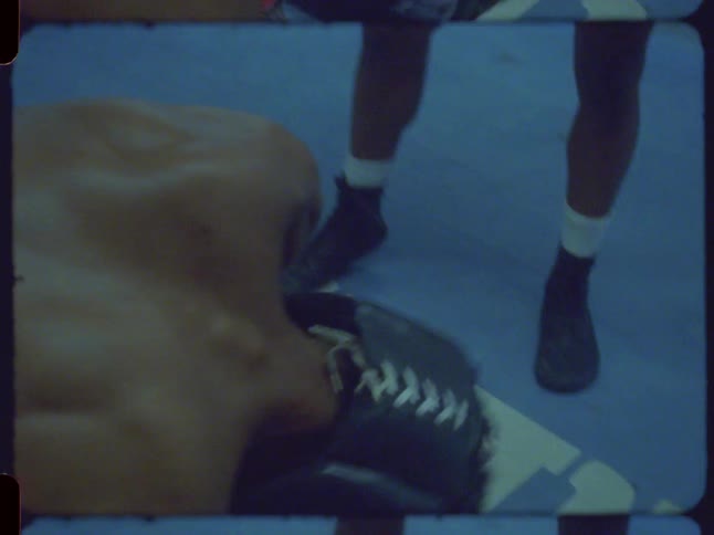 Overhead shot of a boxer wearing headgear doing push-ups on a blue mat in a gym, captured on vintage film