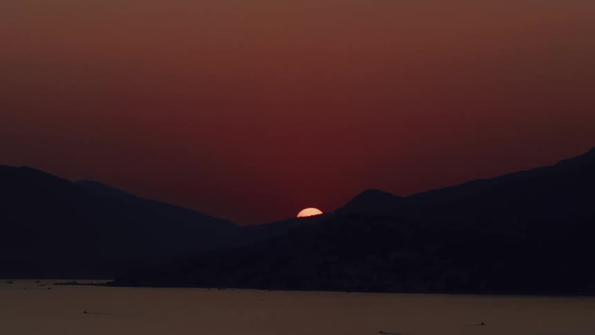 Time-lapse captures the sun setting behind silhouetted mountains, casting a deep red glow across the sky and calm water below