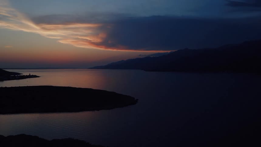 An aerial drone slowly flies over a tranquil sea at dusk, capturing the silhouette of islands against a dramatic sunset sky with vibrant clouds