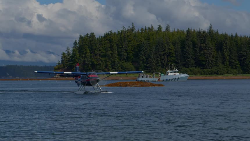 A seaplane taxis on the water in a remote bay with a fishing boat anchored in the background against a forested shoreline