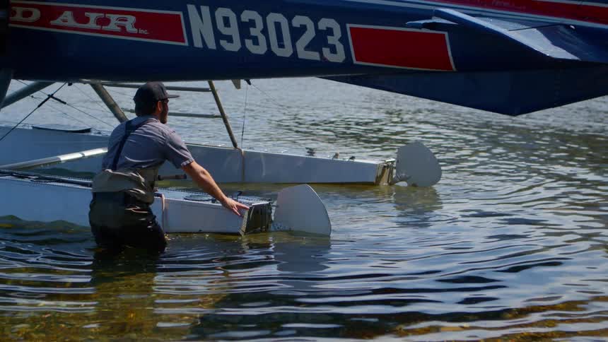 A man in waders holds onto the float of a seaplane as it sits in the shallow water of a lake