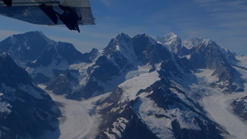 A breathtaking aerial view from an airplane window reveals the vast, snow-covered peaks of a majestic mountain range