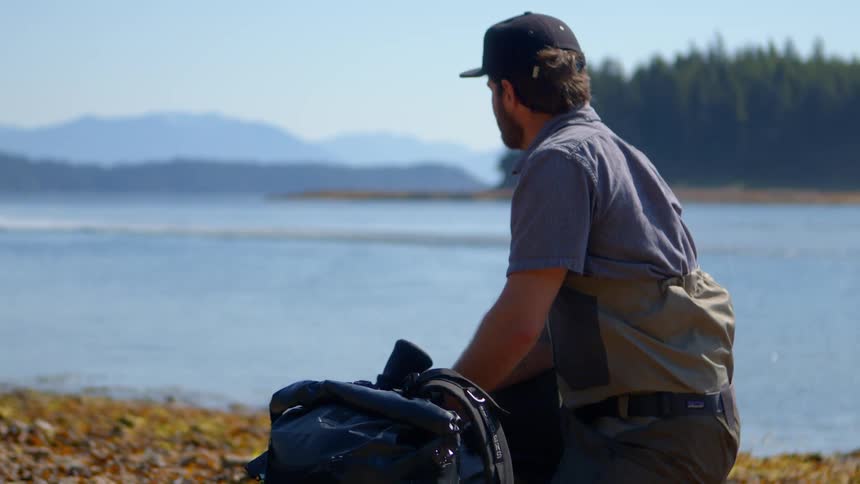 A man in waders unpacks his gear on a remote shoreline while a seaplane taxis away across the bay
