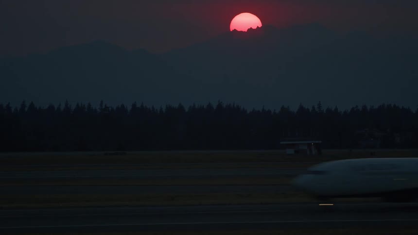 An airplane taxis across a runway at dusk with a dramatic red sun setting behind distant silhouetted mountains