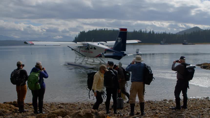 A group of adventurers with backpacks watch and photograph a seaplane as it departs from the shore of a remote lake