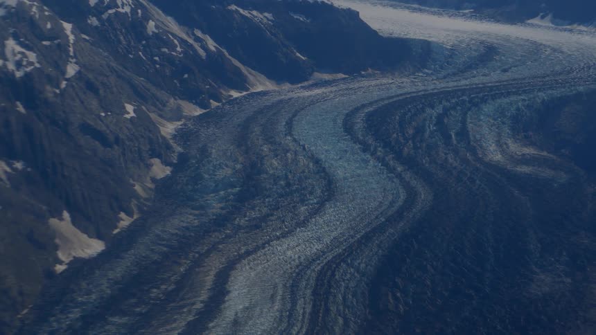 A static aerial view shows a vast, winding glacier flowing through a rugged mountain valley