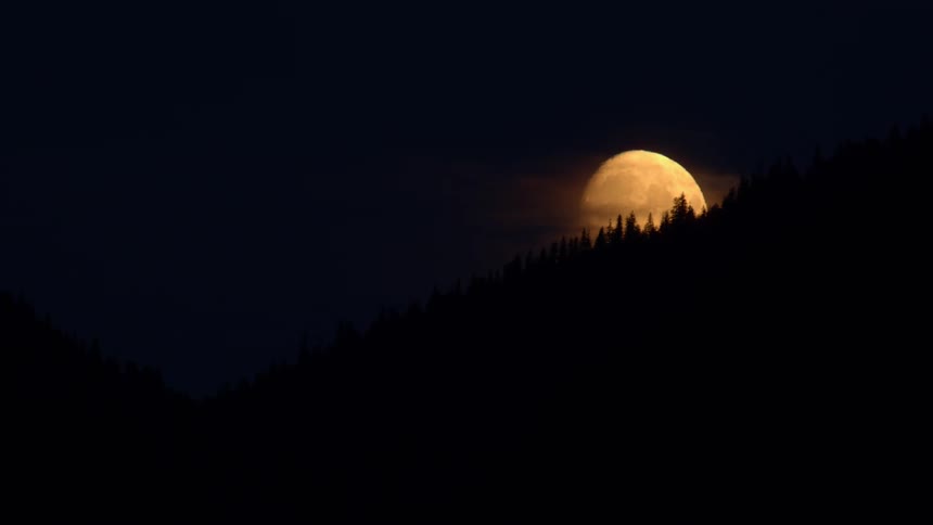 A glowing orange full moon peeks out from behind the silhouette of a mountain forest on a dark night