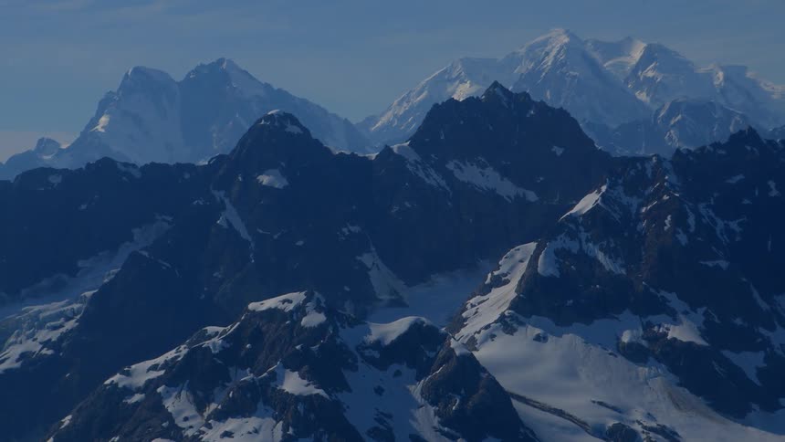 A majestic, snow-capped mountain range stands under a clear blue sky, with the view slowly zooming in on the distant peaks