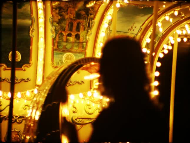 A silhouetted woman contemplates a brightly lit classic carousel spinning at night in this nostalgic, vintage-style footage