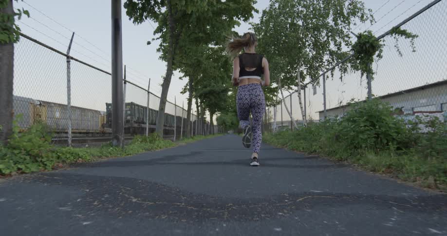 Woman jogs down a road lined with chain link fences near a railroad