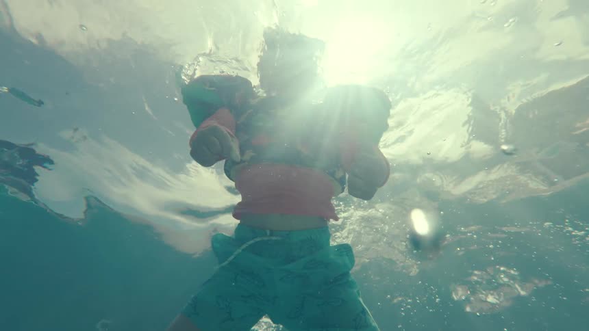 An underwater slow-motion, point-of-view shot shows a child in a snorkel mask and floaties giving a thumbs-up while swimming