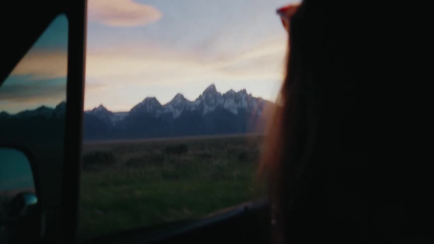 A passenger in a moving car photographs the majestic Grand Teton mountain range at sunset