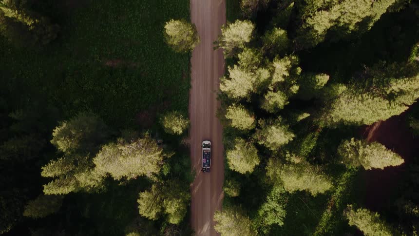 A top-down aerial view follows an Van as it travels along a remote dirt road surrounded by a lush evergreen forest
