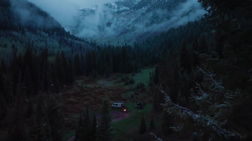 A remote campsite with a van and a warm campfire is nestled in a foggy mountain valley surrounded by a dense evergreen forest at dusk