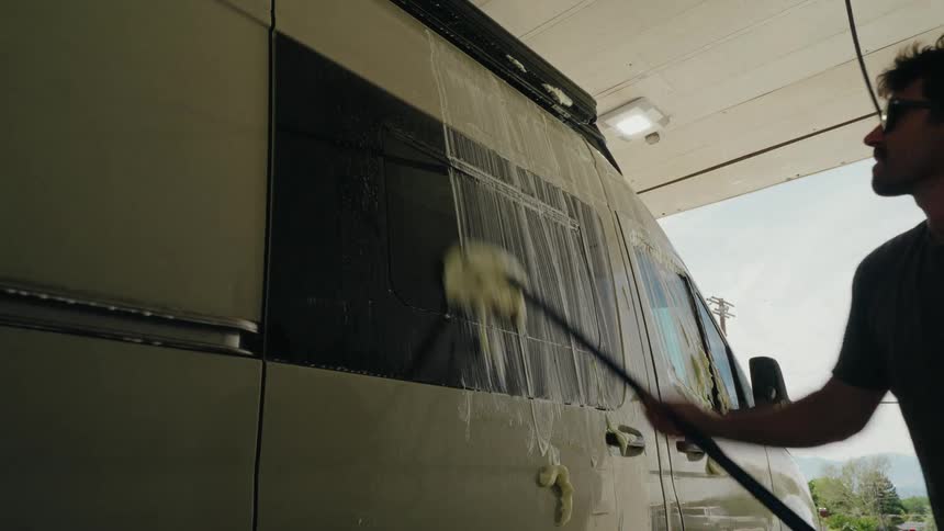 A man in sunglasses diligently washes his camper van with a soapy brush at a self-service car wash
