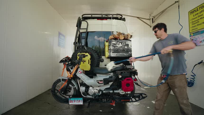 A man diligently washes his fully equipped overlanding camper van and the motorcycle mounted on the back at a self-service car wash