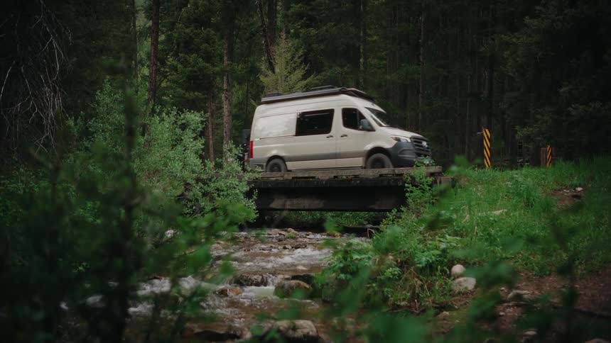 A tan camper van slowly crosses a rustic wooden bridge over a flowing creek in a lush green forest