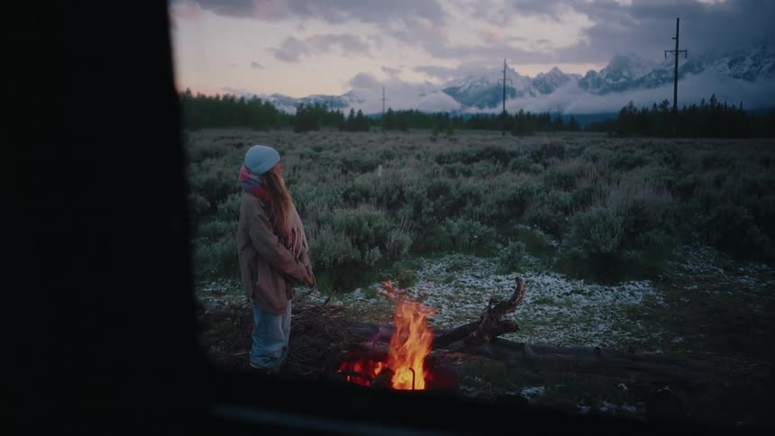 A girl in a beanie and scarf stands by a campfire at twilight, looking out over a snowy meadow towards a majestic mountain range